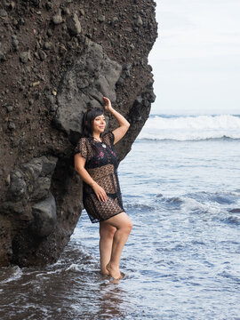 Smiling Woman in Black Lace Outfit Standing in Ocean Waves by Volcanic Cliff