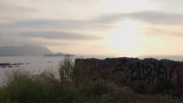 Coastal landscape showing a bay with industrial port structures, distant mountains, and wind turbines on the horizon at sunset