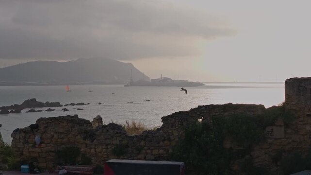 Sailboats navigating calm waters near a historic stone wall, showing industrial facilities and mountains under an overcast sky