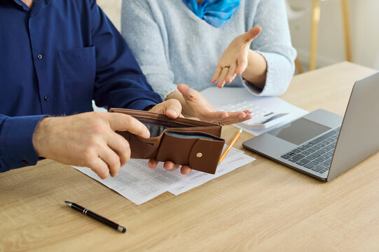 Seniors couple budgeting with empty wallet and bills. Closeup hands review debt at laptop, calculating monthly costs and limited pension income. Concept shows financial stress in retirement.