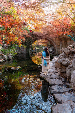 Solo female tourist looking at traditional Chinese stone bridge creating a circular moon bridge reflection in stream surrounded by red and golden autumn trees
