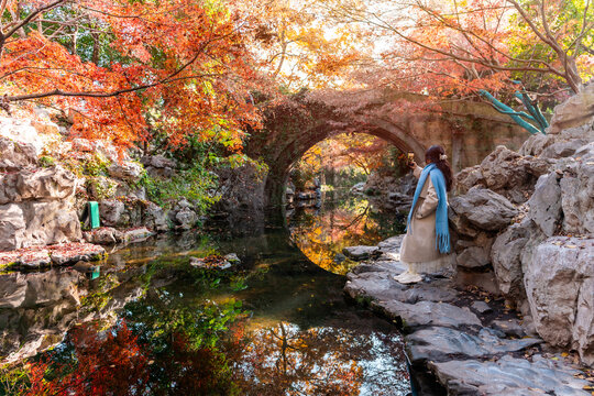 Solo female tourist taking a photo of traditional Chinese stone bridge creating a circular moon bridge reflection in stream surrounded by red and golden autumn trees
