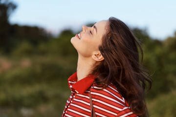 Obraz premium Young brunette woman looking up at the sky with a gentle smile outdoors. She wears a casual striped shirt capturing a peaceful lifestyle moment in the golden sunlight.