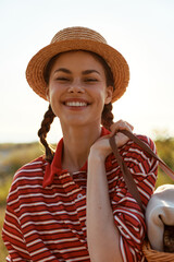 Obraz premium Young woman with braided hair wearing a straw hat and striped shirt smiles while carrying a picnic basket outdoors during a golden hour sunset in a peaceful nature spot.