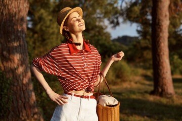 Obraz premium Young woman enjoying summer lifestyle outdoors in a garden. She wears a striped shirt, straw hat, and carries a wicker basket in the warm sun. Concept of leisure, vacation, and nature.