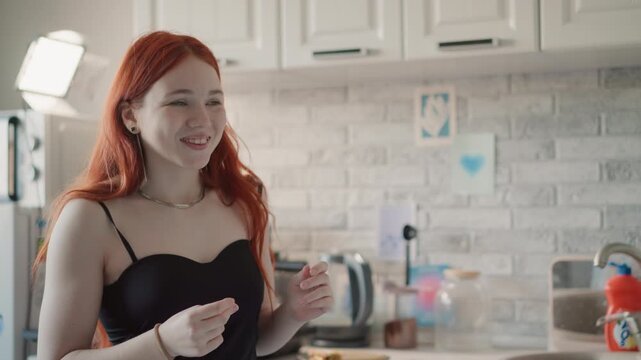 Redheaded woman speaking in kitchen sharing prediction and playful foresight, casual black dress, necklace and freckles, warm sunlight and candid smile, conversational gesture toward camera