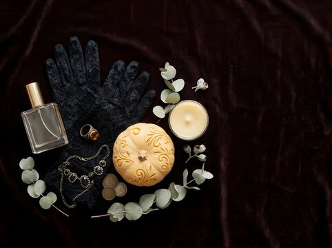 Flat lay composition of a luxury perfume bottle, black lace gloves, an intricately carved pumpkin, vintage jewelry, and a lit candle arranged on a dark brown velvet fabric background.