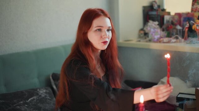 White redhead holds lit candle while performing divination ritual in cozy room, smoke drifting past colorful altar and trinkets, focused clairvoyant expression and slow hand movements creating moody,