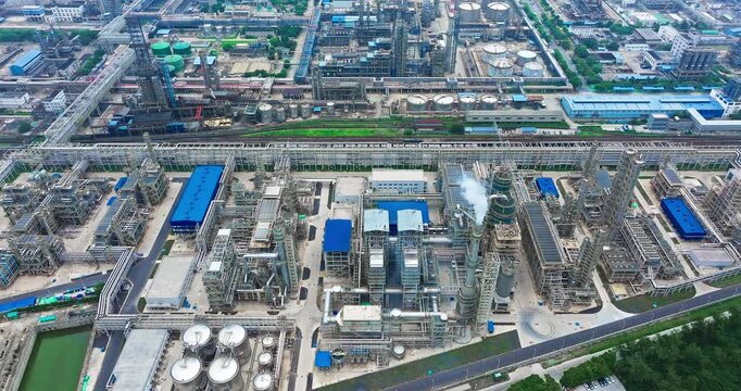 Industrial petrochemical refinery with distillation towers, storage tanks, pipe racks, process equipment, and service roads viewed from above in daylight.