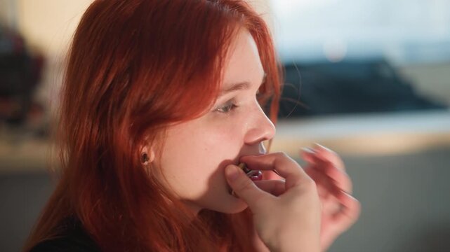 Bright caucasian redhead biting fortune cookie in sunlit kitchen, fingers holding fragile pastry, surprised smile, paper message peeked out, casual lifestyle vibe and playful curiosity