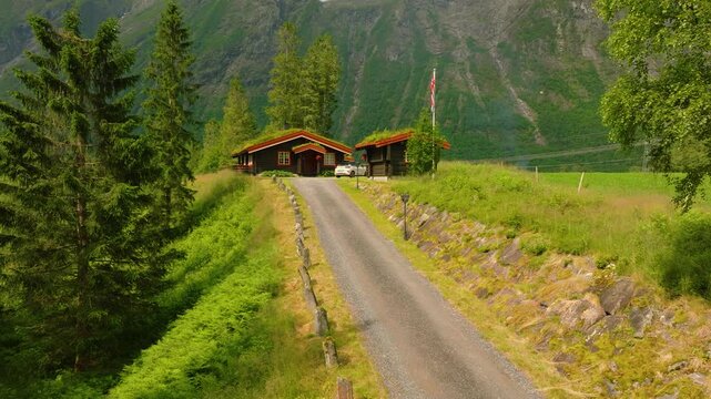 Smooth forward drone flight approaching a traditional Norwegian wooden cabin in a lush mountain valley near Trollstigen. The cabin is surrounded by forest and meadows, with steep green mountains 