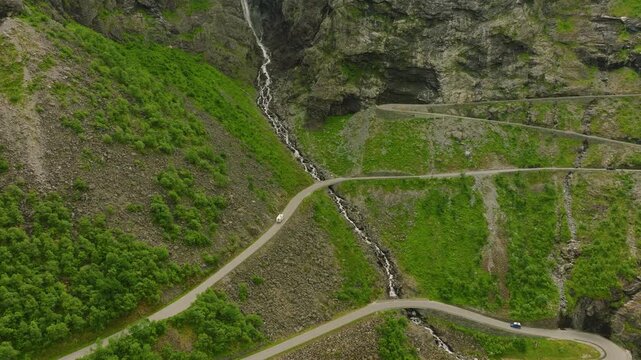 Aerial drone footage of Trollstigen, one of Norway&rsquo;s most spectacular mountain roads. The winding road climbs through a dramatic rocky mountain landscape with steep cliffs, green vegetation, waterfall