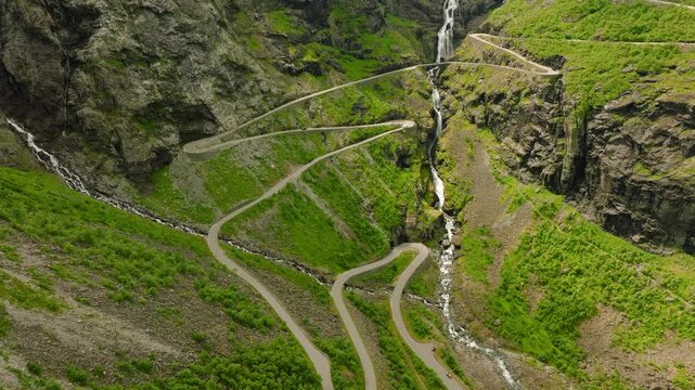 Aerial drone footage of Trollstigen, one of Norway&rsquo;s most spectacular mountain roads. The winding road climbs through a dramatic rocky mountain landscape with steep cliffs, green vegetation, waterfall