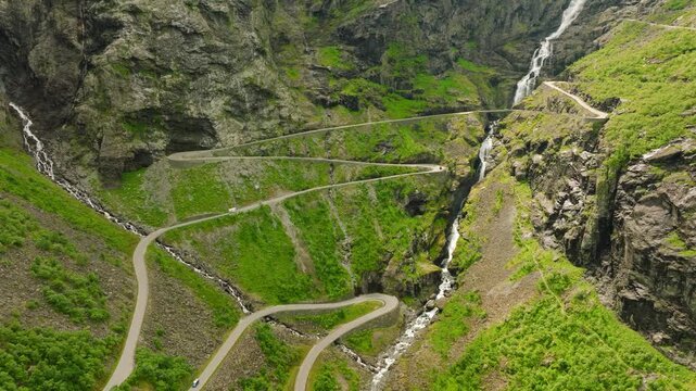 Aerial drone footage of Trollstigen, one of Norway&rsquo;s most spectacular mountain roads. The winding road climbs through a dramatic rocky mountain landscape with steep cliffs, green vegetation, waterfall