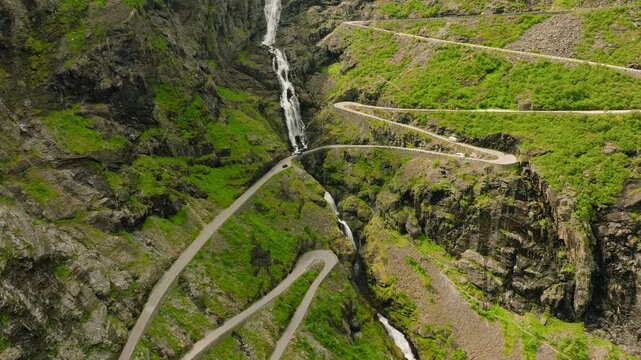 Aerial drone footage of Trollstigen, one of Norway&rsquo;s most spectacular mountain roads. The winding road climbs through a dramatic rocky mountain landscape with steep cliffs, green vegetation, waterfall