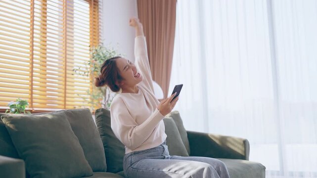 Smiling pretty young Asian woman sitting on cozy couch, using funny mobile apps in living room. Woman at Home, Doing Online Shopping, Messaging Friends, Posting on Social Media