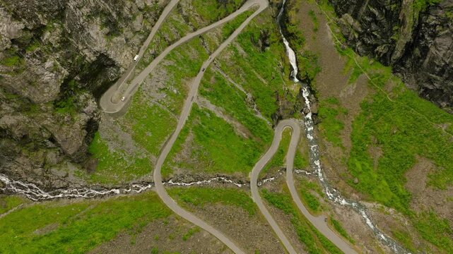 Aerial drone footage of Trollstigen, one of Norway&rsquo;s most spectacular mountain roads. The winding road climbs through a dramatic rocky mountain landscape with steep cliffs, green vegetation, waterfall