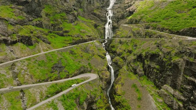 Aerial drone footage of Trollstigen, one of Norway&rsquo;s most spectacular mountain roads. The winding road climbs through a dramatic rocky mountain landscape with steep cliffs, green vegetation, waterfall