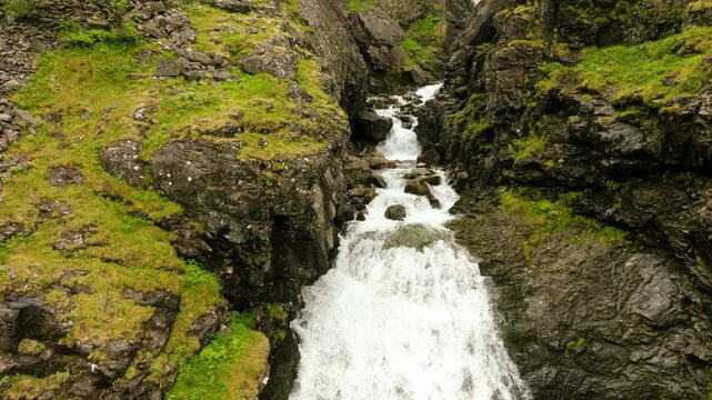 Cinematic drone footage flying over the powerful Stigfossen waterfall at Trollstigen in Norway and passing beneath the historic stone arch bridge spanning the gorge. Dramatic rocky cliffs, rushing 