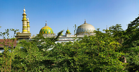 Beautiful mosque domes and minarets with colorful geometric patterns framed by green trees against a clear blue sky © adi