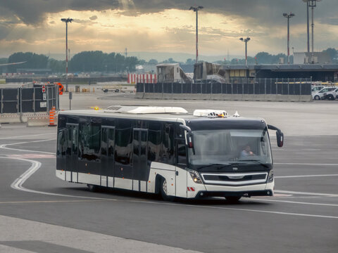 Montreal, Quebec, Canada. August 9, 2026. Side three quarter view of an airport shuttle bus driving on the tarmac under a cloudy sunset sky.