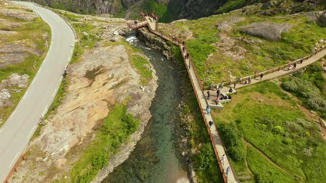 Smooth cinematic drone footage of a scenic viewpoint near Trollstigen in Norway, showing the elevated pedestrian walkway winding above a mountain stream and rocky alpine terrain. Tourists explore 