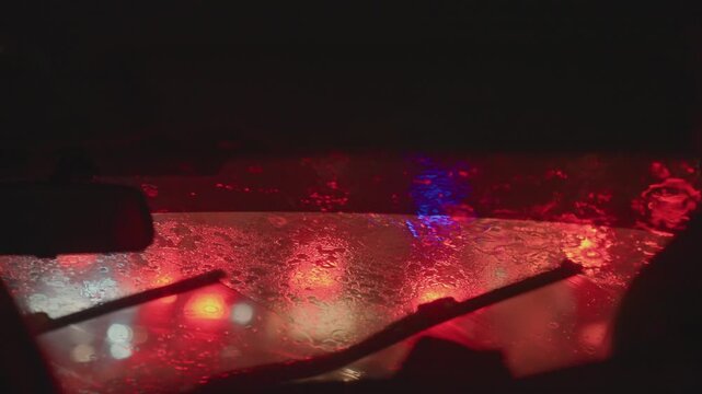 Rain drenched rear window glowing red, blurred brake lights and flashing blue bokeh, windshield wiper scraping droplets, congested city traffic, reflective glass and moody tension
