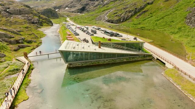 Smooth cinematic drone footage of the Trollstigen Visitor Centre on the Trollstigen mountain plateau in Norway. The modern glass-and-stone building is surrounded by mountain terrain, a clear stream