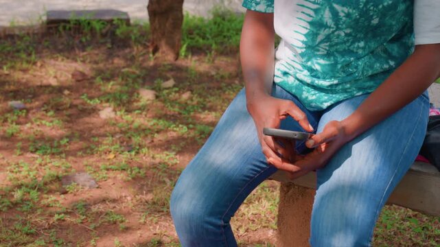 Adult seated on bench holding smartphone, cropped torso and hands visible, denim jeans and green patterned top, sunlight patches on grass, casual moment of scrolling messages, quiet urban park