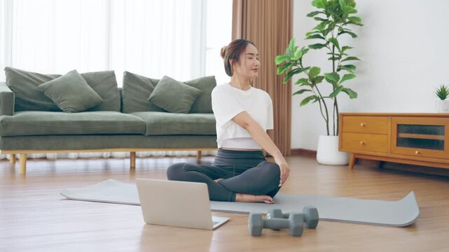 Happy young Asian woman practicing yoga and shoulder stretching at home sitting on floor in living room