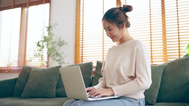 Young Asian woman working from home. Freelancer businesswoman using laptop at sunny room. Student female learning and working at home