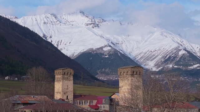 telephoto aerial orbit showcasing two iconic Svan towers in Mestia with snow-covered mountain peaks creating a spectacular alpine backdrop.