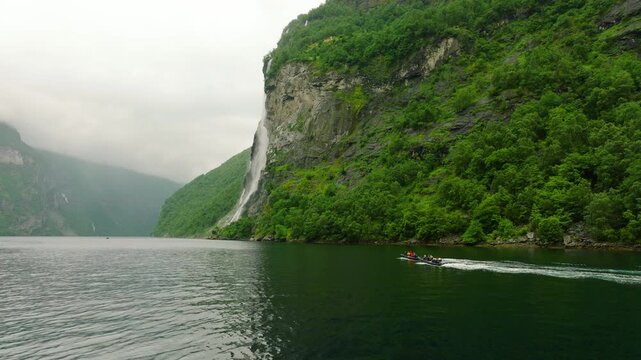Cinematic drone footage flying low over the waters of Geirangerfjord, showcasing the spectacular Seven Sisters waterfall cascading down the steep green cliffs. Small sightseeing boats travel across th