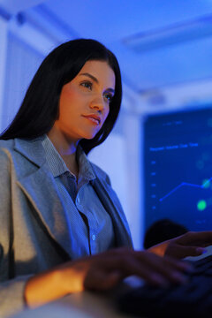 Focused female cybersecurity analyst monitoring computer systems at night in a blue-lit office, representing network security, threat detection, data protection, and digital defense.