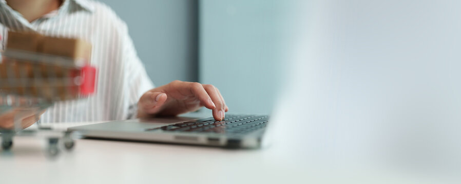 Woman shopping online on a laptop beside a miniature cart with packages, representing e-commerce, product browsing, online purchase, digital retail, delivery, payment, and consumer convenience.