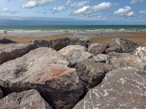 Rocky breakwater and sandy beach with distant swimmers under a bright summer sky in Wissant, France