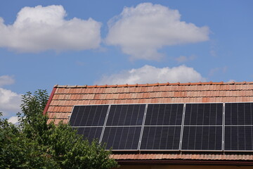 Fototapeta premium Solar panels on a red tiled roof beside a green fruit tree under a blue summer sky with white cumulus clouds