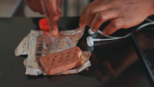 Mother preparing childs snack on countertop, closeup of hands unwrapping chocolate wafer sandwich from foil wrapper beside earbuds and smartphone on glossy surface, warm kitchen light, intimate caring