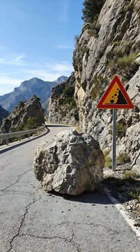 Large gray boulder blocking a narrow mountain road with a yellow triangular rockfall warning sign on the side of the rocky cliff face