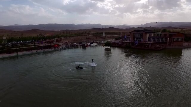 Wide aerial perspective captures multiple flyboard users enjoying water activity on large lake with boats and park facilities visible around.