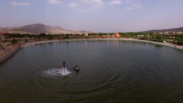 Aerial drone footage captures thrilling flyboard rider soaring above lake surface creating large water splashes and jets with scenic park and mountains in background under clear sky.