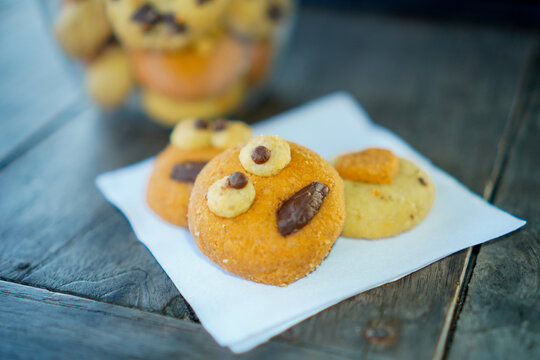 Close-up shot of funny monster face cookies topped with chocolate chips spilling out from a clear plastic jar onto white baking paper and a dark wooden table. Ideal for concepts of creative baking, ki