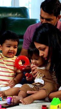 Indian Asian twin infant babies playing with colorful toys while young father and mother sit together on carpet in modern living room at home, capturing joyful family bonding and parenthood moments