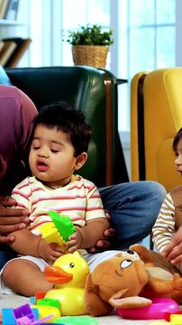Indian Asian twin infant babies smiling and looking at camera while sitting with young parents on carpet surrounded by toys in modern home living room, capturing joyful family portrait and bonding