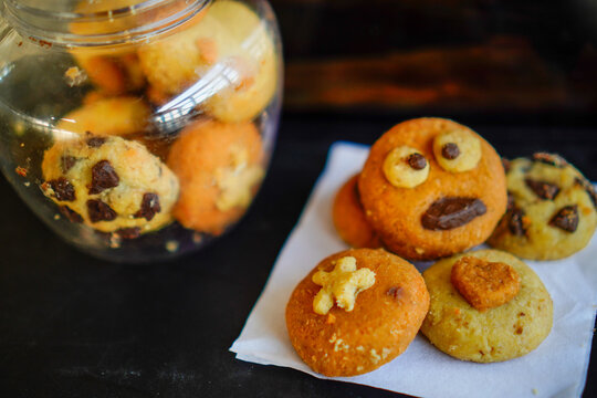 Close-up shot of creative handmade cookies with fun monster face, star, and heart toppings served on white baking paper next to a clear plastic cookie jar. Ideal for concepts of home baking, kids part