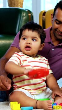 Indian Asian twin infant babies smiling and looking at camera while sitting with young parents on carpet surrounded by toys in modern home living room, capturing joyful family portrait and bonding