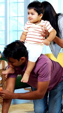 Indian Asian young father giving horse ride to baby boy sitting on his back while mother holds twin infants together in modern home living room, capturing playful family bonding joyful parenting time