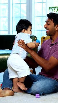 Indian Asian young parents sitting back to back on carpet holding twin infant babies high in air with joyful smiles while babies laugh and enjoy playful bonding time in modern home interior