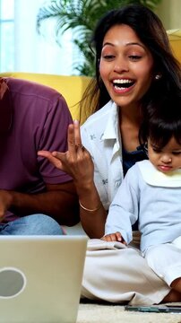 Indian Asian young parents sitting on carpet with twin infant babies attending video call on laptop computer in modern home, smiling and waving while connecting with relatives or friends online