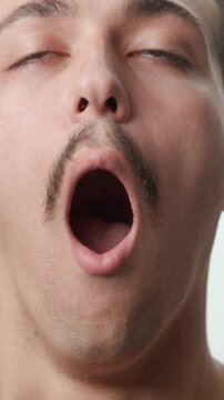 Frontal studio close-up portrait of a tired young Caucasian man with a mustache yawning with eyes closed and looking sleepy on a white background.